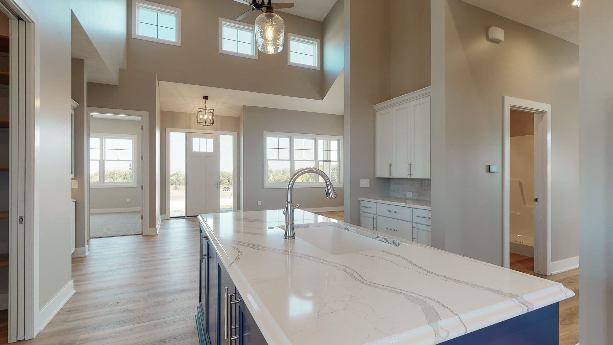 Bright modern kitchen features a large island with a quartz countertop, stainless steel faucet, and blue cabinetry. The open-concept design includes vaulted ceilings, pendant lighting, and hardwood floors leading to other areas of the home.