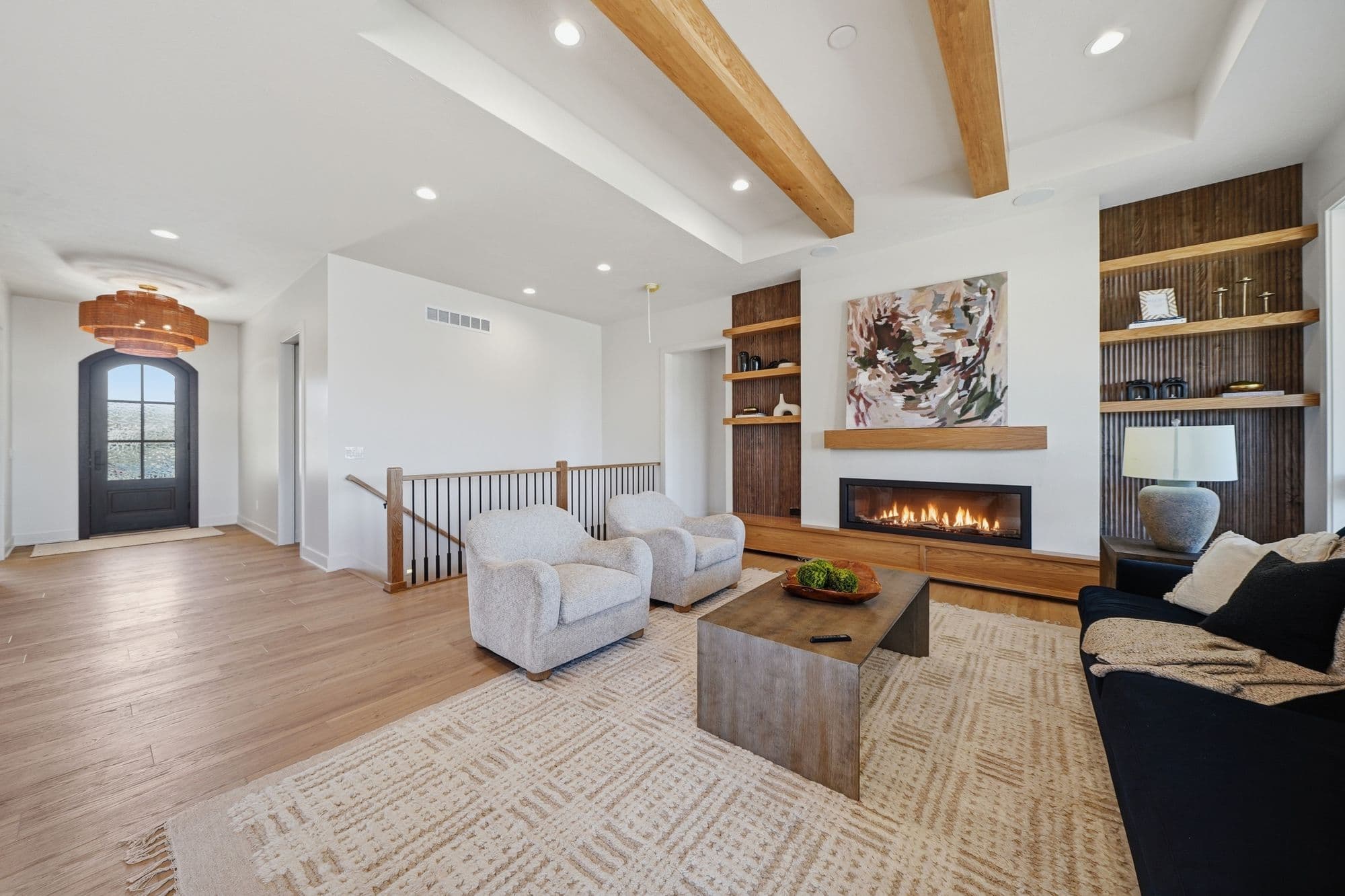Contemporary living room features two armchairs facing a modern fireplace with a wood mantel and abstract art above, with built-in shelving and wood beams on the ceiling. The room is furnished with a coffee table, a dark sofa, and an area rug, all bathed in bright, natural light.